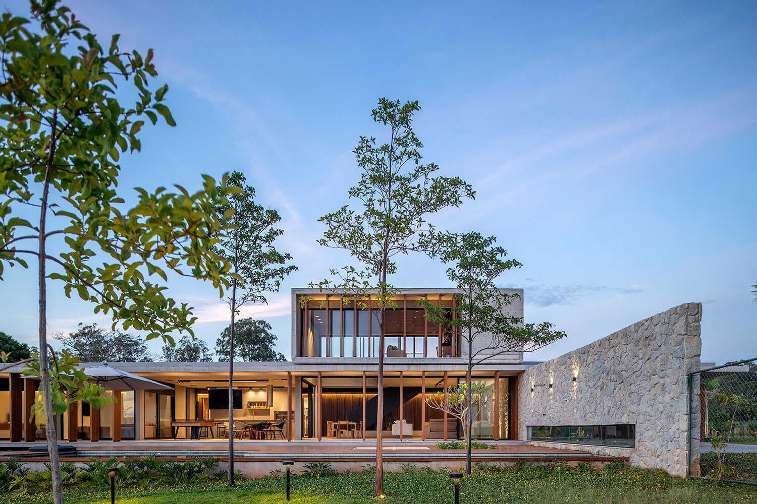 Casa Pedra ao entardecer, com volumes de concreto, madeira e muro de pedra