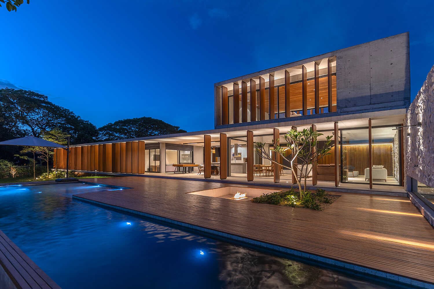 Fachada noturna com piscina, brises de madeira e fire pit